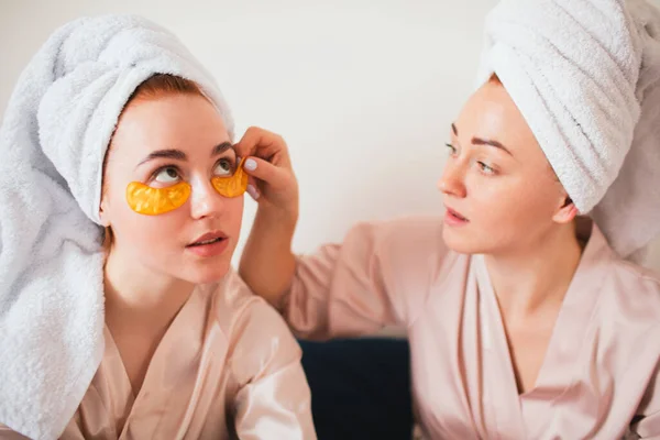 Due giovani donne si divertono con le toppe sotto gli occhi. Due amici in asciugamani e pigiama hanno un divertente festa termale insieme a casa . — Foto Stock