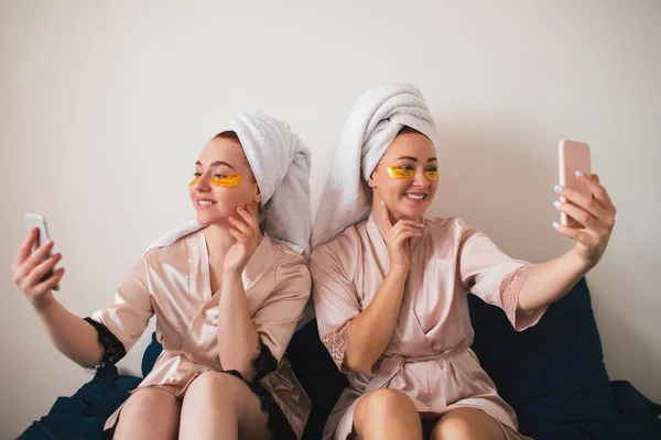 Deux jeunes femmes qui s'amusent avec des patchs sous les yeux. Deux amis en serviettes et pyjamas font une fête spa amusante ensemble à la maison. Prendre des selfies ou utiliser leurs smartphones pour les réseaux sociaux — Photo