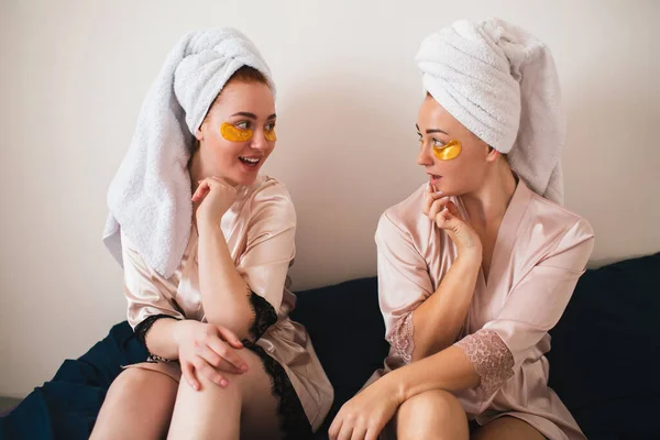 Two young women having fun with patches under their eyes. Two friends in towels and pajamas have a fun spa party together in home. — Stock Photo, Image