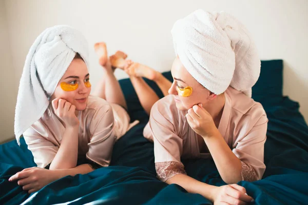 2人の若い女性が目の下にパッチで楽しんでいます。タオルとパジャマを着た2人の友人が家で一緒に楽しいスパパーティーをしています. — ストック写真