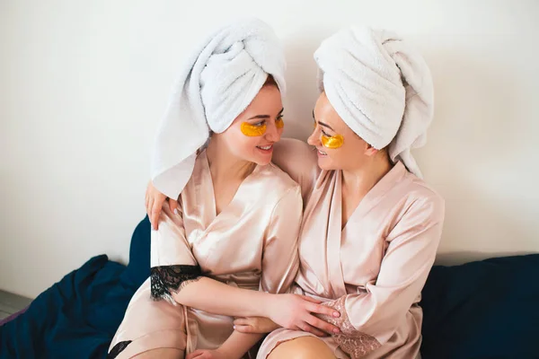 Due giovani donne si divertono con le toppe sotto gli occhi. Due amici in asciugamani e pigiama hanno un divertente festa termale insieme a casa . — Foto Stock