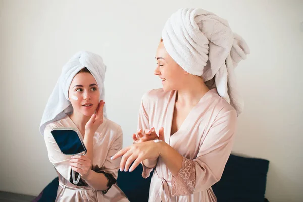 I modelli femminili applicano la crema su mano e faccia. Due giovani donne in asciugamani e pigiama hanno un divertente festa termale insieme a casa . — Foto Stock