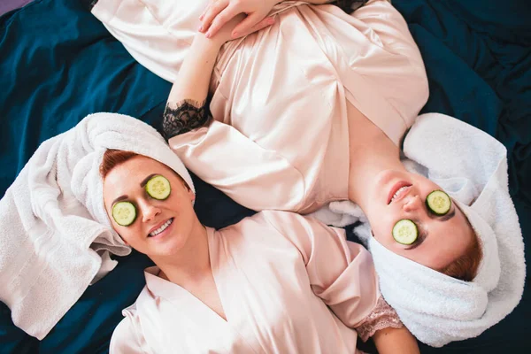 Two female models make an eye mask using fresh green cucumber. Two young women in towels and pajamas have a fun spa party together in home. — Stock Photo, Image