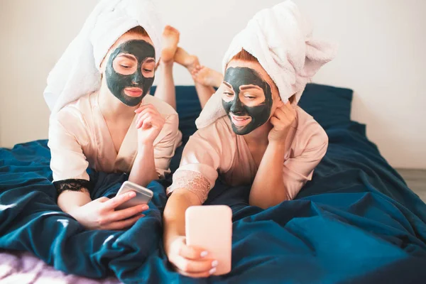 2つの女性モデルは、顔のマスクを作成し、ソーシャルネットワークでの自撮りや通信のためのスマートフォンや電話を使用しています。タオルとパジャマを着た2人の若い女性が家で一緒に楽しいスパパーティーをしています. — ストック写真