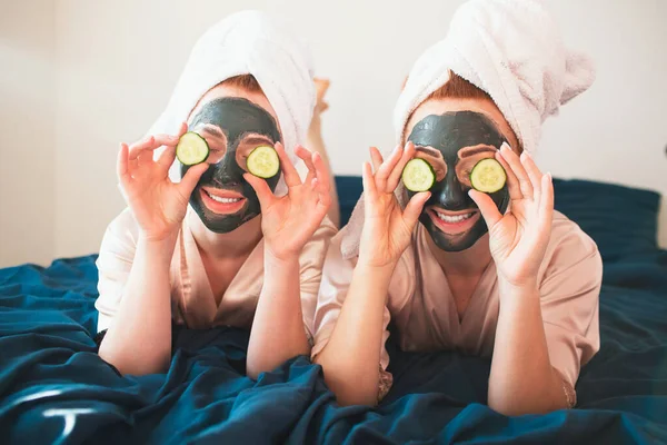 Due modelli femminili fanno la maschera di faccia e usando il cetriolo verde fresco. Due giovani donne in asciugamani e pigiama hanno un divertente festa termale insieme a casa . — Foto Stock