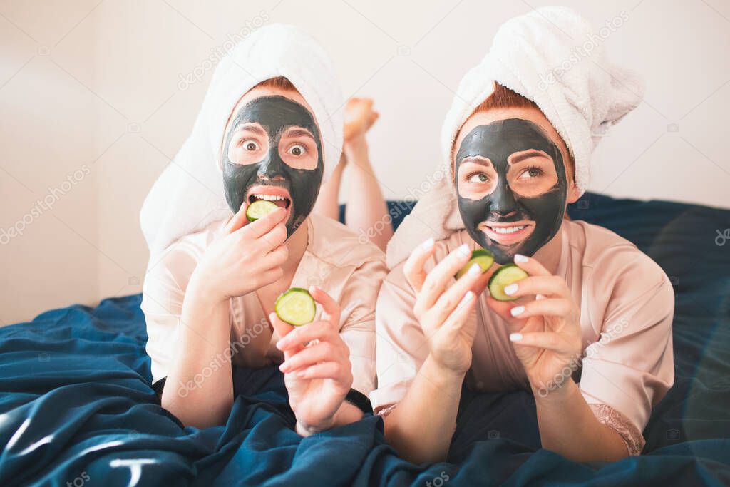 Two female models make face mask and using fresh green cucumber. Two young women in towels and pajamas have a fun spa party together in home.