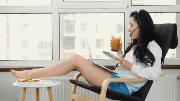 Mladá žena pracuje doma během karantény. Náročné multitasking zaměstnanec call centrum pít džus a usmívat se. Videohovory a konverzace. Diskuse online přes sluchátka. — Stock video