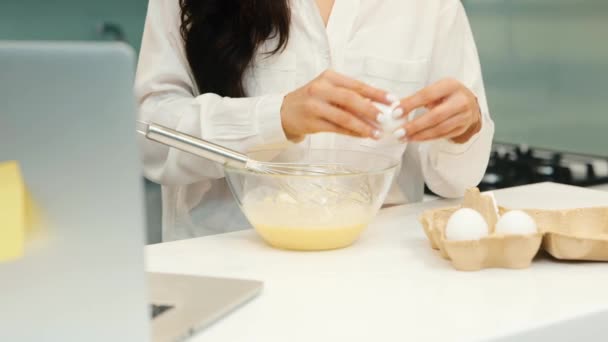 Młoda kobieta w domu podczas kwarantanny. Powolny ruch dodawania jednego jajka i mieszania go w misce. Gotowanie i przygotowywanie śniadania. Wielozadaniowa kobieta. Praca i gotowanie jednocześnie. — Wideo stockowe