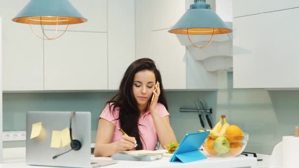 Mujer joven en la cocina durante la cuarentena. Chica hablando por teléfono y tomando notas. Persona multitarea y multifuncional. Mujer trabajadora en cocina y comiendo sándwich . — Vídeos de Stock