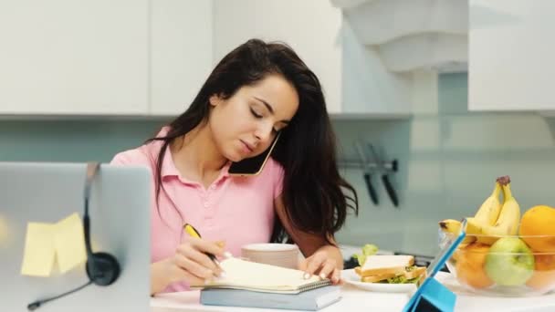 Ung kvinna som arbetar hemma under karantänen. Flickan pratar i telefon och antecknar samtidigt. Multifunktionell multitasking person och anställd. — Stockvideo