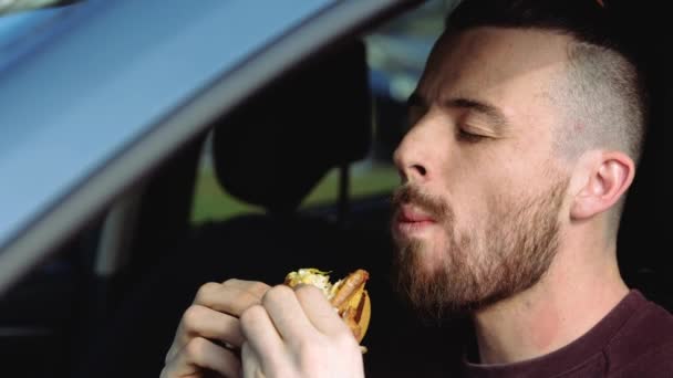Giovanotto in macchina. Siediti e goditi il gustoso delizioso hamburger. Mordilo diverse volte. Godetevi il pasto in macchina. Vista laterale. Rallentatore . — Video Stock