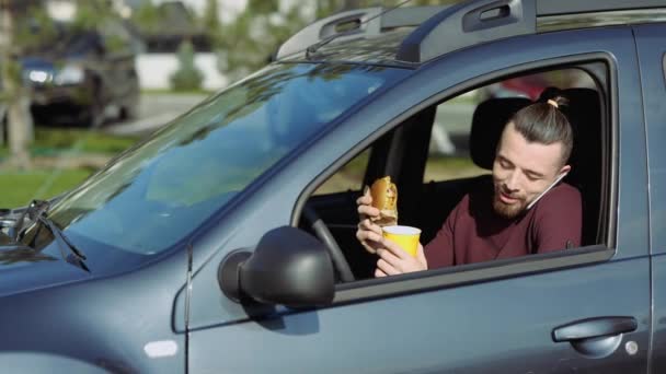 Unge man i bilen. Prata i telefon under lunchtid eller frukost. Äta hamburgare och dricka i bilen. Uttrycksfull hipster. — Stockvideo