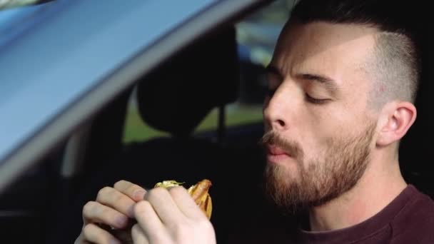 Fiatalember a kocsiban. Közelkép portré hipster eszik ízletes finom hamburger két kézzel. Rágja és élvezi az ételt. Ízletes reggeli vagy ebéd autóban. — Stock videók