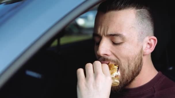 Mladý muž chce auto. Portrét hipsterského chlapa žvýkajícího chutné chutné hamburgery a pijícího z papírového kelímku. Užijte si snídani nebo oběd v autě. — Stock video