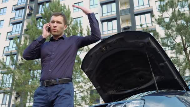 Junger Mann vor dem Auto. Stehen Sie daneben und unterhalten Sie sich emotional auf dem Smartphone. Kaputtes Auto mit Motor funktioniert nicht. Geöffnete Motorhaube. Hilfe durch Telefonanruf. — Stockvideo