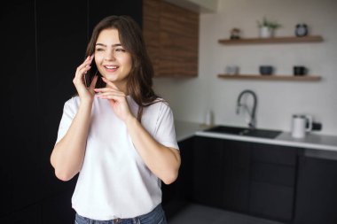 Karantina sırasında mutfakta genç bir kadın. Yalnız dur ve telefonda konuş. Çevrimiçi kablosuz iletişim. Uzaktan kumandalı ev işi. İş kadını..