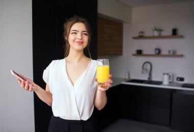 Karantina sırasında mutfakta genç bir kadın. Kız elinde portakal suyu ile telefonu ve bardağı tut. Olumlu neşeli iş kadını gülümsemesi. Merkez ofiste uzaktan çalışma.