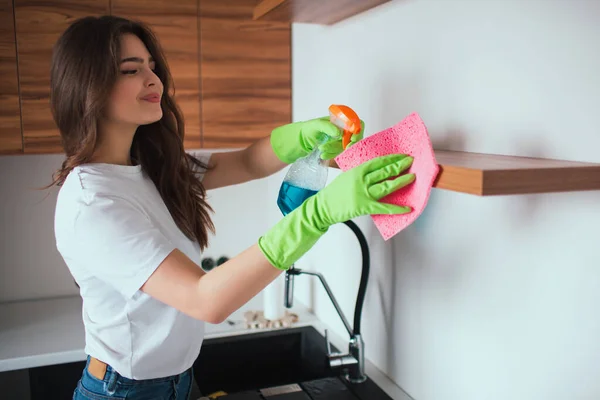 Young woman in kitchen during quarantine. Girl using spray for cleaning shelves from dust. Household and care. Attractive positive girl smile. — Stock Photo, Image