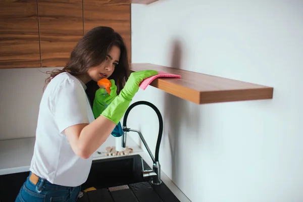 Молода жінка на кухні під час карантину. Дівчина ретельно прибирає поверхню полиці. Використовуючи тканину та розпилювач для чищення. Носіть зелені рукавички навколо рук . — стокове фото
