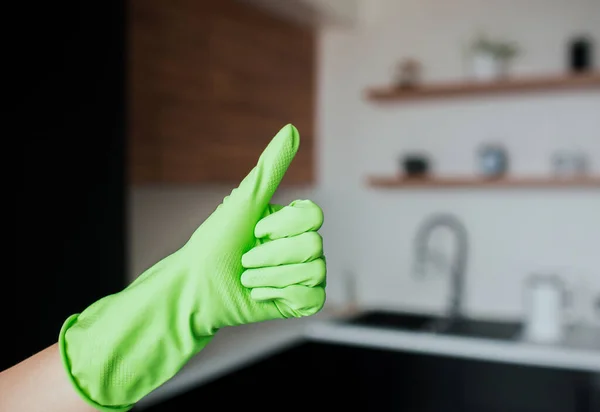 Young woman in kitchen during quarantine. Female hand in green glove hold big thumb up. Kitchen interior with blurred background. — Stock Photo, Image