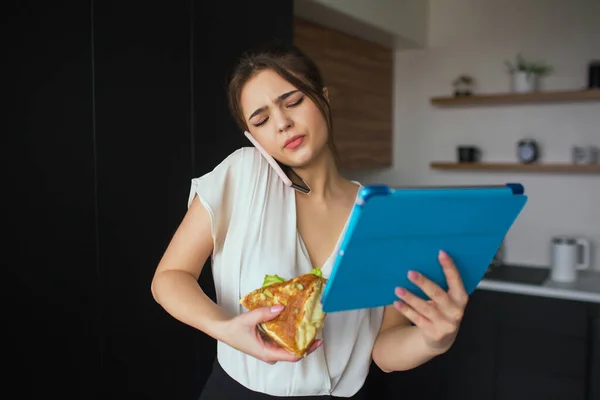 Ung kvinna i köket under karantänen. Affärskvinna pratar i telefon. Håll stor tablett och smörgås i händerna. Hemmakontor och distansarbete. Lunchtid. — Stockfoto