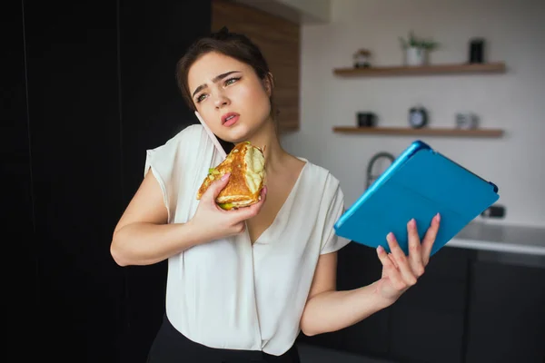 Ung kvinna i köket under karantänen. Hon pratar i telefon och äter smörgås. Affärskvinna håller blå tablett i handen. Hemmakontor och distansarbete. Kommunikation och online-samtal. — Stockfoto