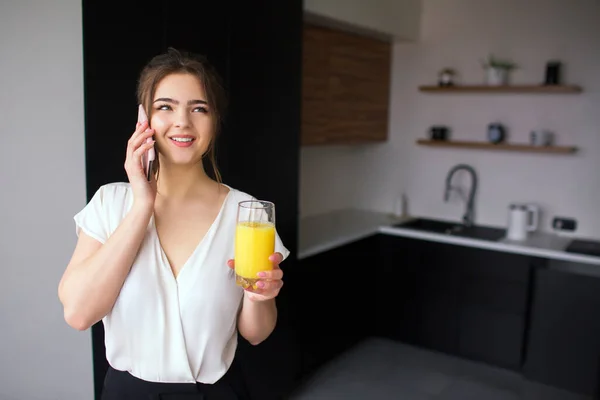 Ung kvinna i köket under karantänen. Flickan pratar i telefon och ler. Affärskvinna håller i ett glas apelsinjuice. Trådlöst samtal online. — Stockfoto