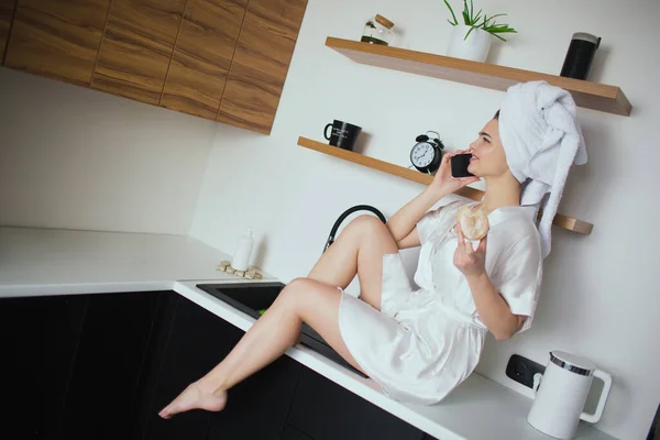 Jeune femme en cuisine pendant la quarantaine. Jolie fille s'asseoir et parler au téléphone après la douche Tenez beignet dans la main. Sexy fille passer du temps dans la cuisine . — Photo