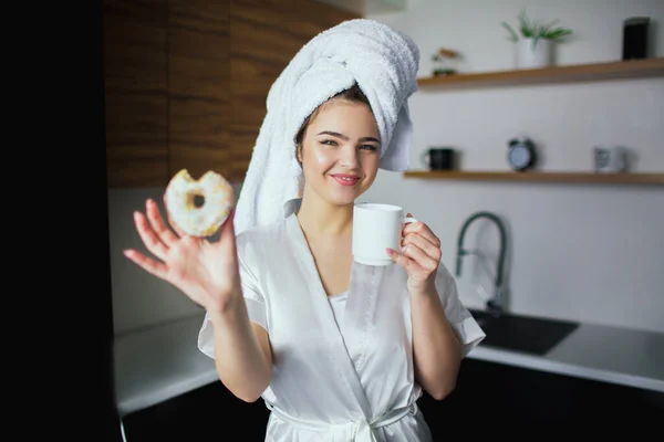 Young woman in kitchen during quarantine. Lovely beautiful girl after bathing posing on camera in white towel and robe. Hold white cup and tasty donut. — Stock Photo, Image