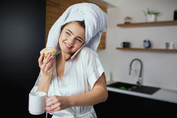 Young woman in kitchen during quarantine. Talking on phone and smile. Breakfast after bathing. Woman in white robe and towel eat donut. — Stock Photo, Image