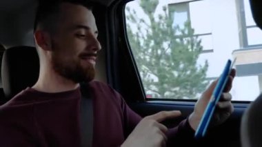 Arabanın içindeki genç adam. Adam arka koltukta oturup oyun oynuyor ya da tablet kullanarak internette sörf yapıyor. Yalnız iyi eğlenceler. Mutluluktan gülümsüyor. Taksiye ya da özel arabaya binmek..