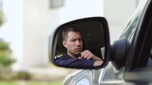 Νεαρός μέσα στο αυτοκίνητο. Μια άποψη από τον πλευρικό καθρέφτη του επιχειρηματία καυχιέται σκληρά και βαθιά. Καλύψτε το στόμα με το χέρι. Μόνος στο αυτοκίνητο. Θολή φόντο. — Αρχείο Βίντεο