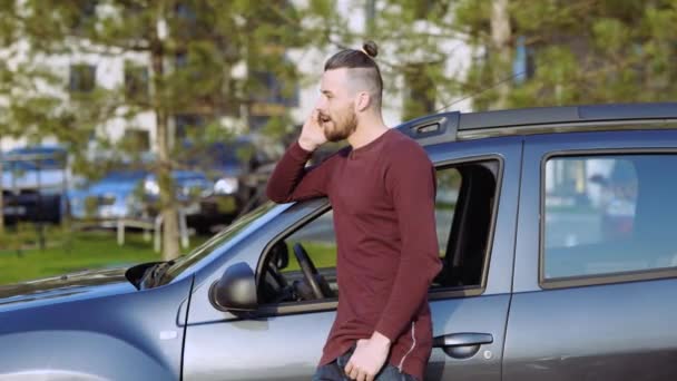 Een jongeman buiten de auto. Hipster aan de telefoon. Vrolijke positieve persoon te wachten naast de auto op straat. Gesprek. Mooi zonnig weer buiten. — Stockvideo