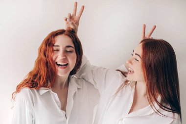 İki kızıl saçlı kız kardeş, geniş gömleklerle beyaz bir arka planda izole bir şekilde duruyorlar. İki genç kadın kamerada oynaşıyor.