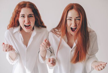 İki genç kadın sinirlenip kameraya bağırdı. Öfke, saldırganlık, öfke hissettiler. İki kızıl saçlı kız kardeş, geniş gömleklerle beyaz bir arka planda izole bir şekilde duruyorlar.