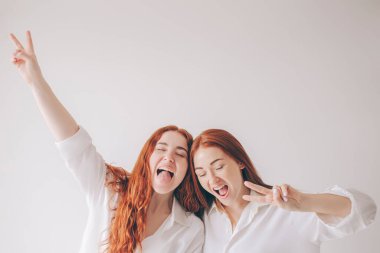 İki kızıl saçlı kız kardeş, geniş gömleklerle beyaz bir arka planda izole bir şekilde duruyorlar. İki genç kadın kamerada oynaşıyor. Surat asıp eğleniyorlar.