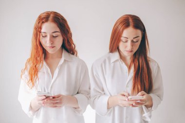 İki kızıl saçlı kız kardeş, geniş gömleklerle beyaz bir arka planda izole bir şekilde duruyorlar. İki genç kadın sosyal ağlara bağımlıdır. Bütün zamanlarını akıllı telefonlarda geçirirler.