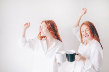 İki kızıl saçlı kız kardeş, geniş gömleklerle beyaz bir arka planda izole bir şekilde duruyorlar. İki genç kız kahve ya da çay içip eğleniyorlar.