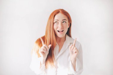 Kızıl saçlı kadın geniş gömleklerle beyaz bir arka planda tek başına duruyor. Kız işaret parmaklarıyla yukarı işaret ediyor.