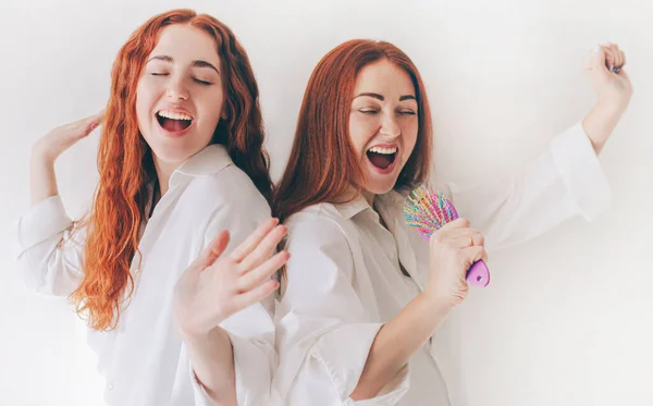 Two young women sing in makeshift microphones - hairbrushes. Two red-haired sisters stand isolated on a white background in spacious oversized shirts.