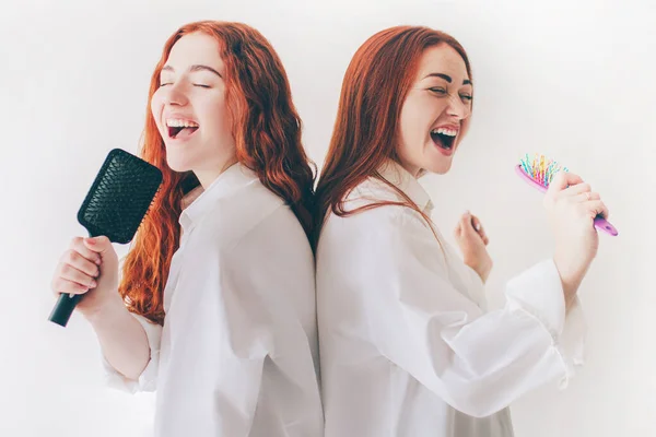 Two red-haired sisters stand isolated on a white background in spacious oversized shirts.