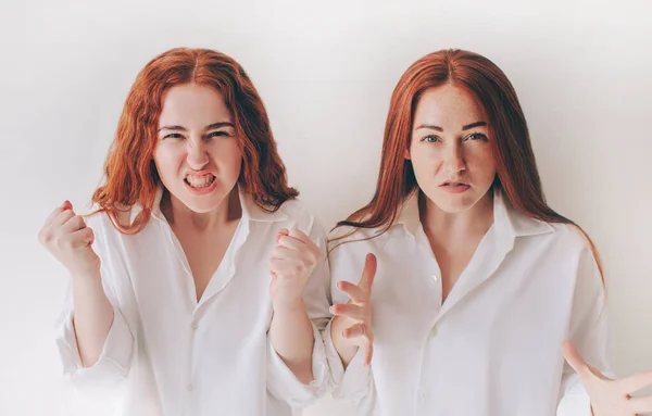 Two young women got angry and shout at the camera They feel rage, aggression, anger. Two red-haired sisters stand isolated on a white background in spacious oversized shirts