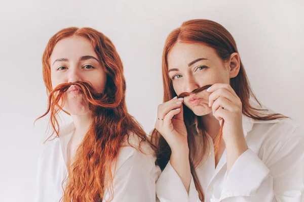 Two red-haired sisters stand isolated on a white background in spacious oversized shirts. Two young women fooling around on camera. They make faces and have fun — Stock Photo, Image