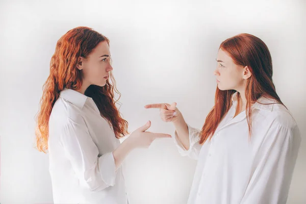 Two red-haired sisters stand isolated on a white background in spacious oversized shirts. TWO young girls accuse each other of their deeds.