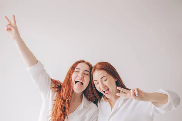 Two red-haired sisters stand isolated on a white background in spacious oversized shirts. Two young women fooling around on camera. They make faces and have fun — Stock Photo, Image