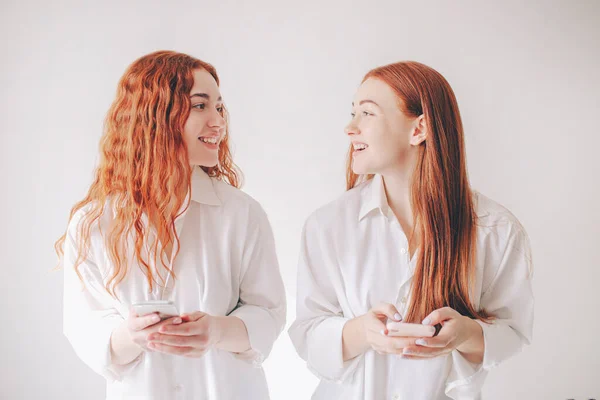 Deux sœurs aux cheveux roux se tiennent isolées sur un fond blanc dans des chemises surdimensionnées spacieuses. Deux jeunes femmes accro aux réseaux sociaux passent tout leur temps sur un smartphone — Photo