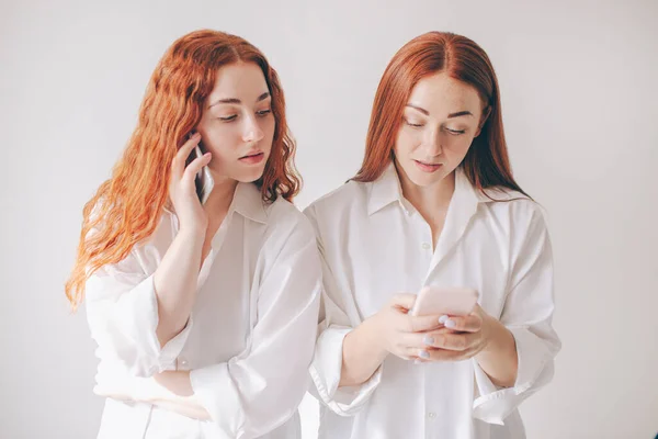 Two red-haired sisters stand isolated on a white background in spacious oversized shirts. Two young women are addicted to social networks spend all their time in a smartphone