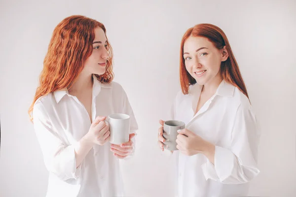 Kaksi punatukkaista siskoa seisoo valkoisella taustalla tilavissa ylimitoitetuissa paidoissa. Kaksi nuorta tyttöä juoda kahvia tai teetä ja on ystävällinen chat . — kuvapankkivalokuva