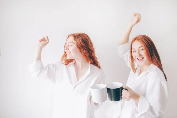 Two red-haired sisters stand isolated on a white background in spacious oversized shirts. Two young girls drink coffee or tea and fool around and have fun — Stock Photo, Image
