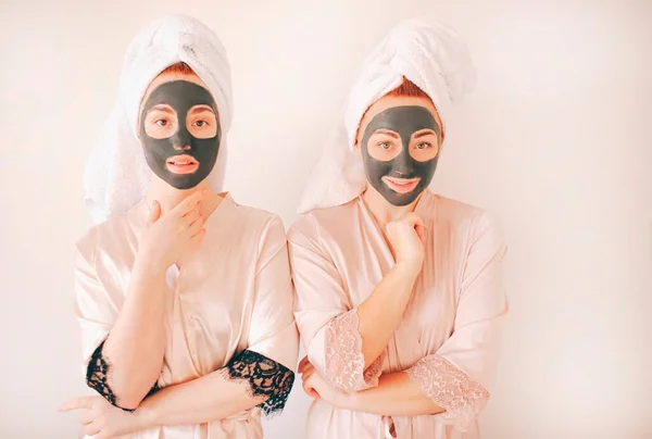 Due giovani sorelle in accappatoi con gli asciugamani in testa isolate su uno sfondo bianco. Spa a casa. Fanno una maschera per la cura della pelle sul viso. E applicato sulla pelle del viso . — Foto Stock